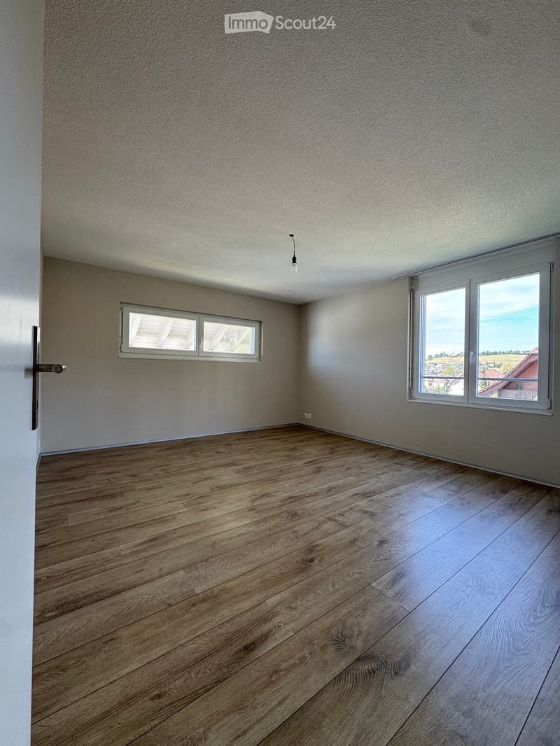 Empty room with wooden flooring, light grey walls, and two windows offering a view of the outside.