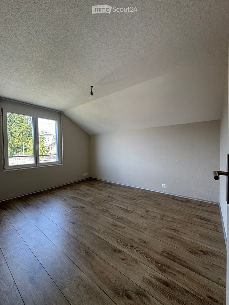 Empty room with wooden flooring, white walls, and a large window letting in natural light. The room has a simple, modern design with a single light fixture on the ceiling.