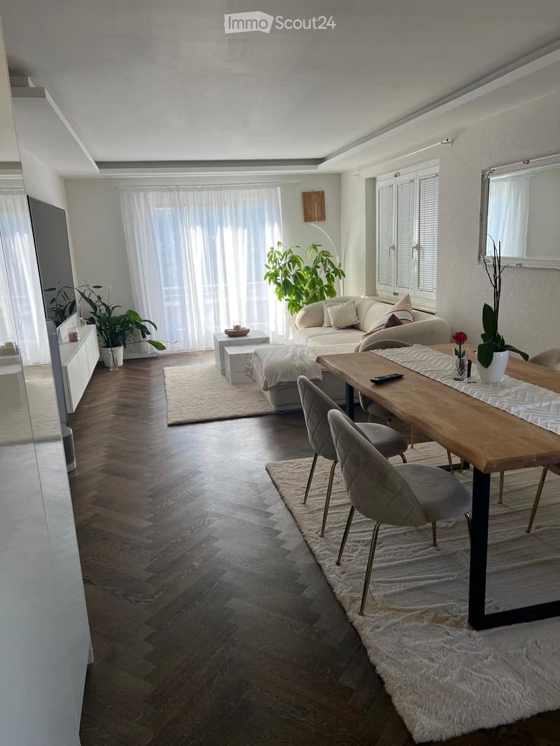 Modern living and dining area with a sofa, dining table, chairs, plants, and a large mirror. The space features wooden flooring, large windows with white curtains, and a cozy rug.