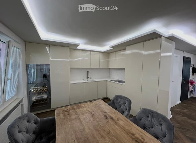 Modern kitchen with light-colored cabinets, a wooden dining table, two grey upholstered chairs, a stainless steel sink, and a built-in refrigerator.