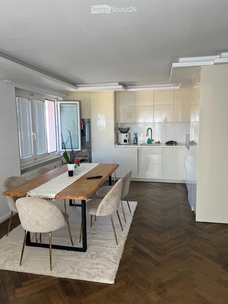 A modern, open-plan dining and kitchen area with a wooden dining table, six chairs, a white rug, and a sleek kitchen with white cabinets and stainless steel appliances.