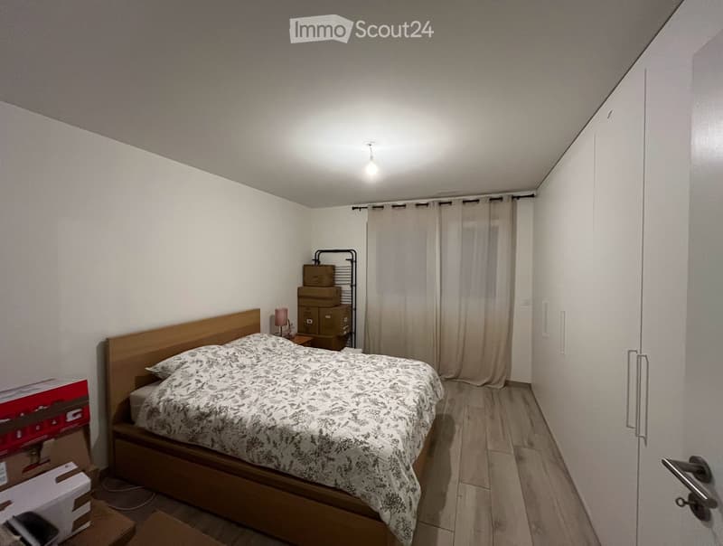 Bedroom with a wooden bed, nightstand, lamp, and curtains. Cardboard boxes and a rack are visible in the background.