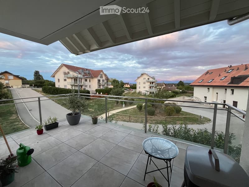 Balcony with tiled floor, metal railing, small table and chair, potted plants, and a view of a residential area with houses and greenery.