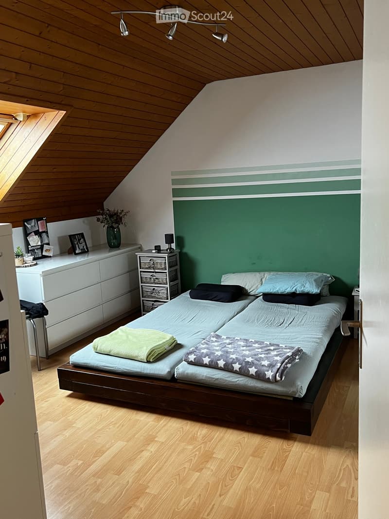 Cozy attic bedroom with wooden slanted ceiling, light wooden floor, double bed with two mattresses, nightstand, drawer unit, and decorative items.