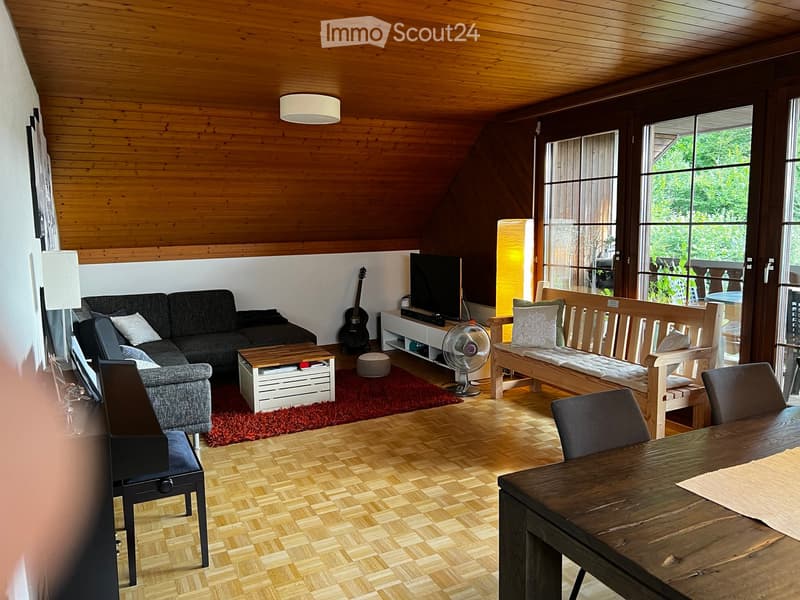 Bright living room with wooden ceiling and herringbone flooring, featuring a grey sectional sofa, coffee table, guitar, fan, and dining area with a wooden table and chairs.