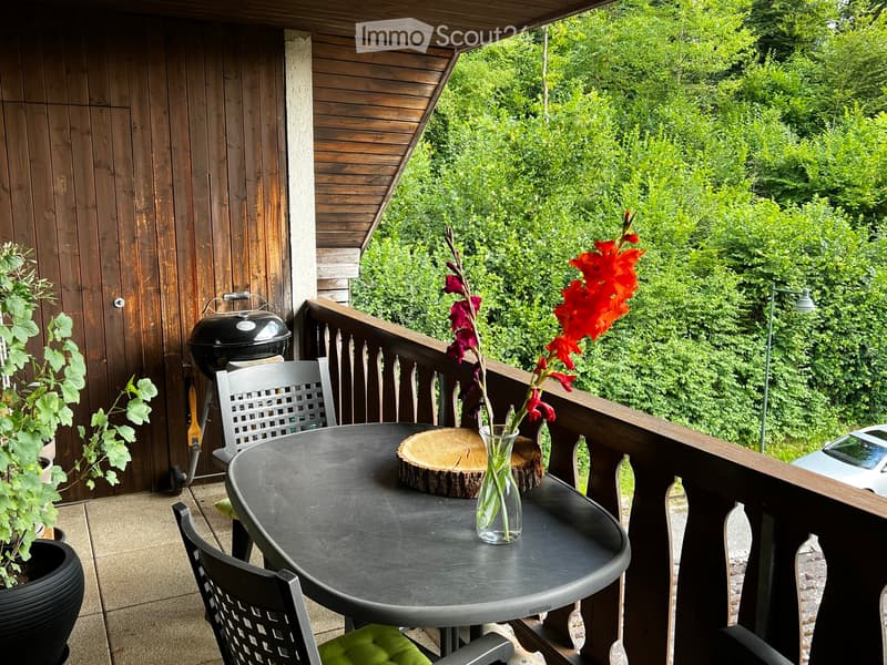 Balcony with a round table, two chairs, a vase with red flowers, and a grill, surrounded by greenery.