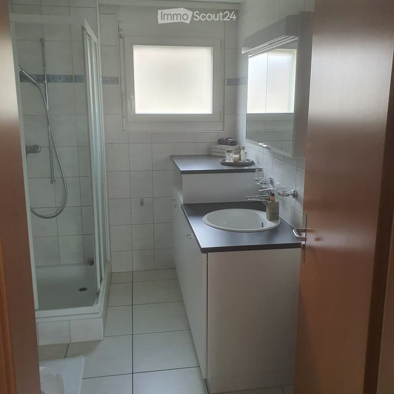 Modern bathroom with white tiles, a sink with a faucet, a mirror, a shower area, and a small cabinet.