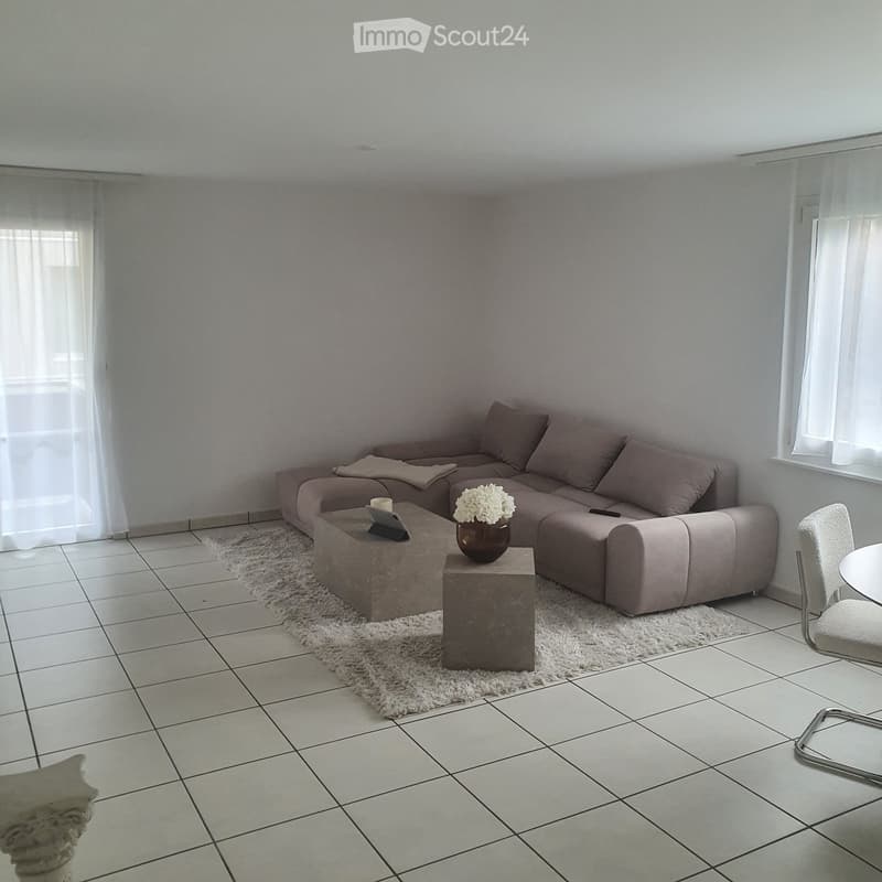 Modern living room with a grey sectional sofa, coffee table, small round table, and decorative vase with flowers. The room features tiled flooring and large windows with sheer curtains.