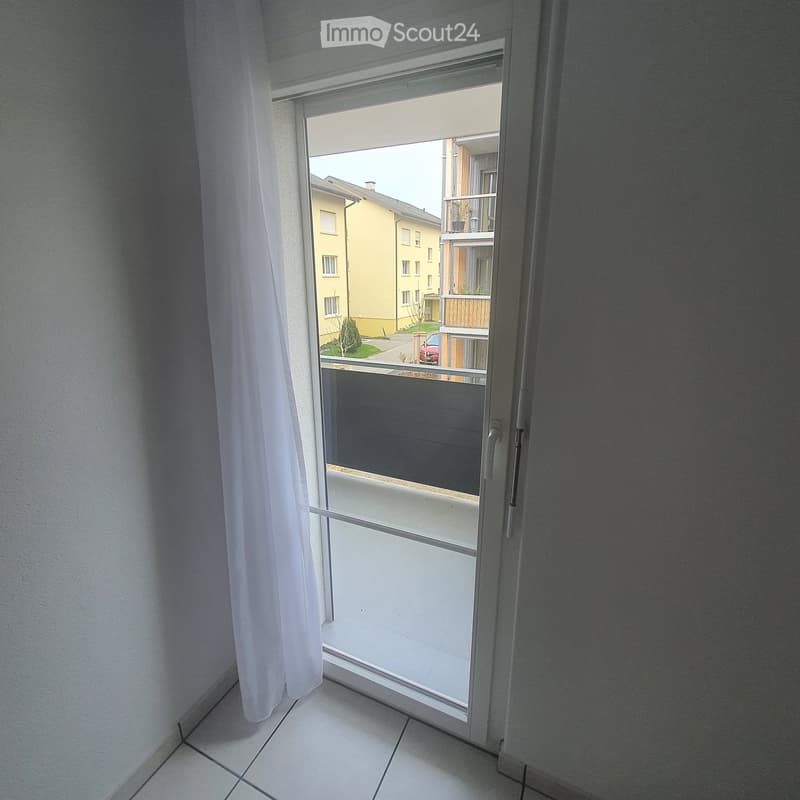 View from a room through an open sliding door onto a balcony, showing a residential area with buildings and a parked car.