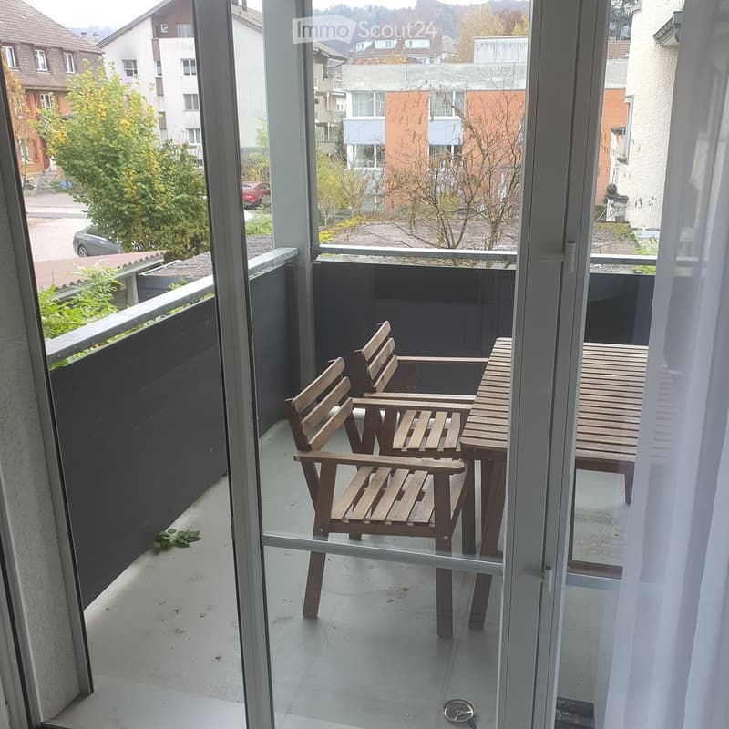 Balcony with two wooden chairs and a small table, overlooking a residential area with houses and trees.