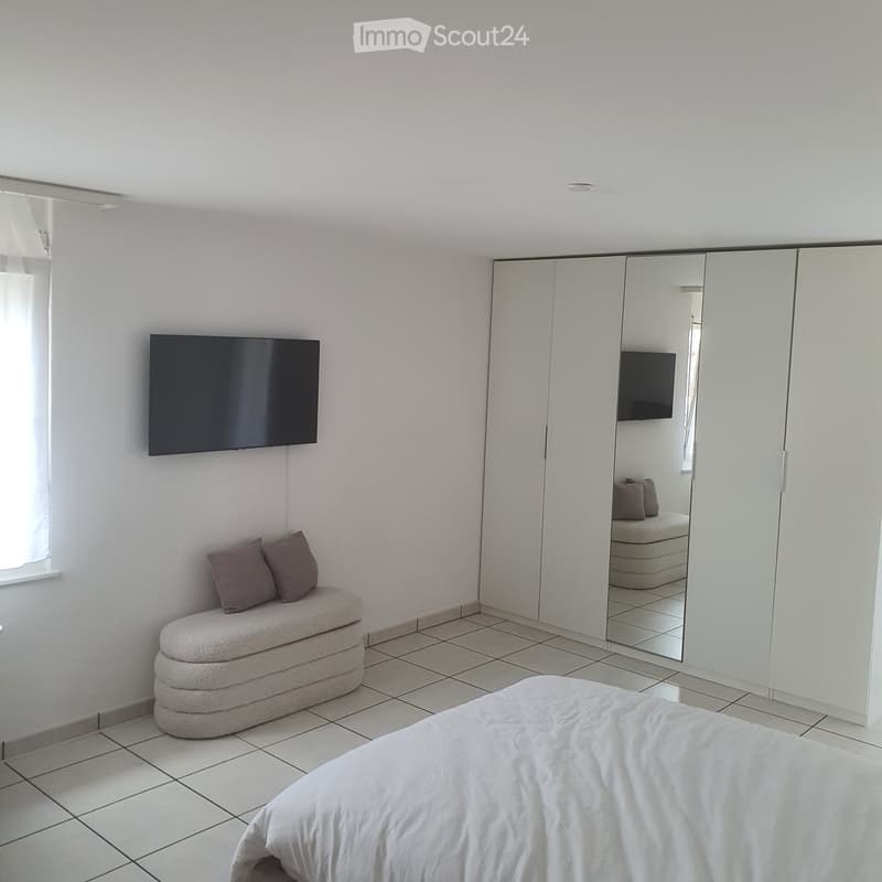Modern bedroom with a white bed, beige bench, two purple pillows, a large mirror wardrobe, and a wall-mounted TV.