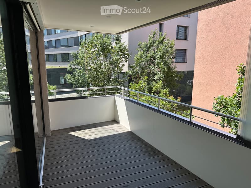 Modern balcony with wooden decking, white walls, metal railing, and a view of greenery and neighboring buildings.