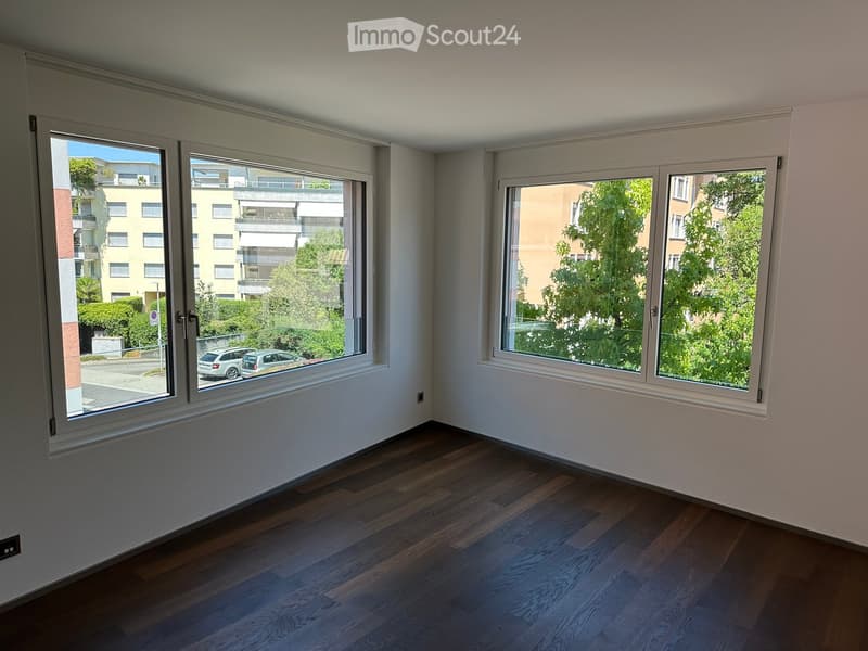 Empty room with wooden flooring and large windows offering views of the street and greenery outside.