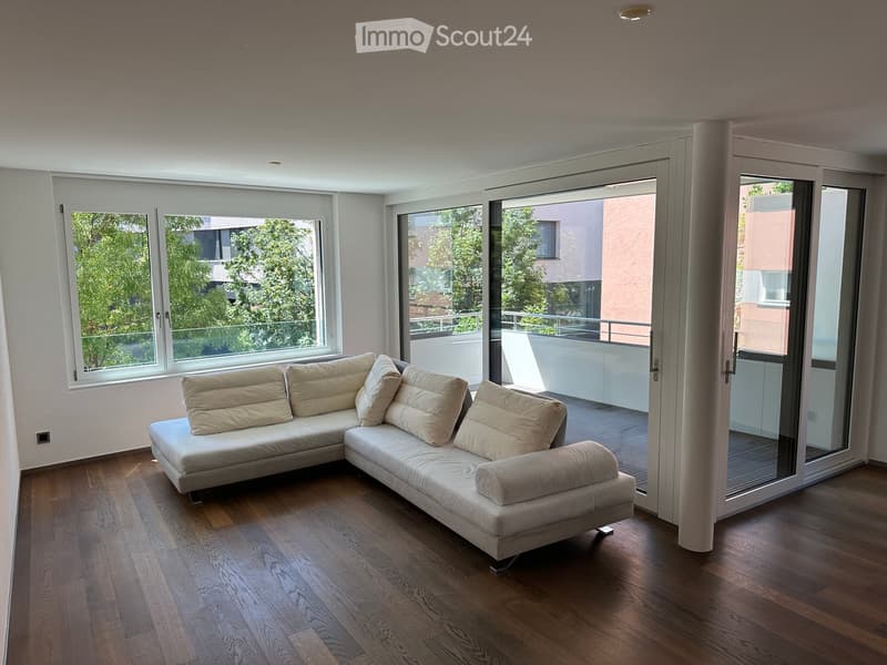 Modern living room with a large sectional sofa, wooden flooring, and large windows offering a view of greenery. A sliding glass door leads to a balcony.