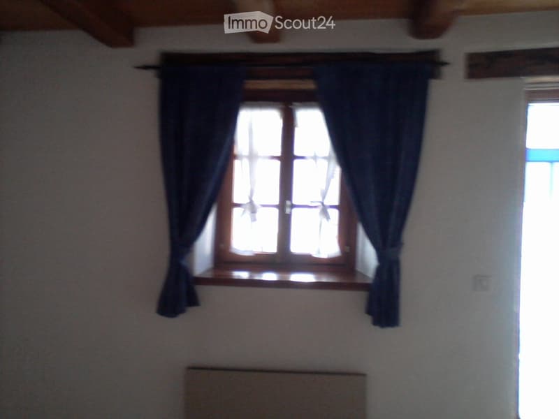Empty room with wooden ceiling beams, a window with dark curtains, and a radiator below the window.