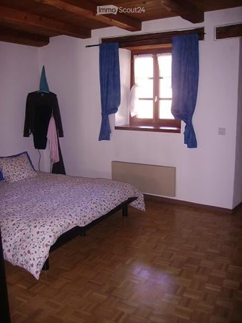 Cozy bedroom with wooden flooring, a bed with floral bedding, a window with blue curtains, a radiator, and a wardrobe with hanging clothes.