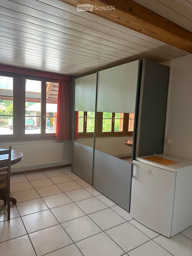 Modern kitchen with white cabinets, tiled floor, and a small round table with a chair. There is a refrigerator and a window with red curtains.