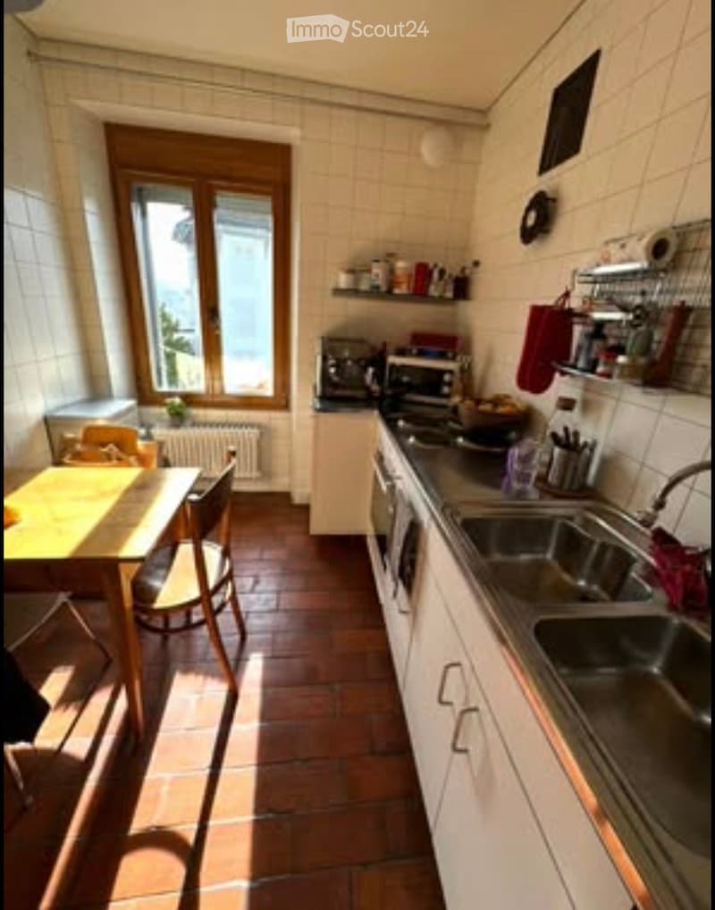 A well-lit kitchen with white cabinets, a stainless steel sink, and a small dining area with a table and chairs. The floor is tiled, and there are various appliances and utensils visible.