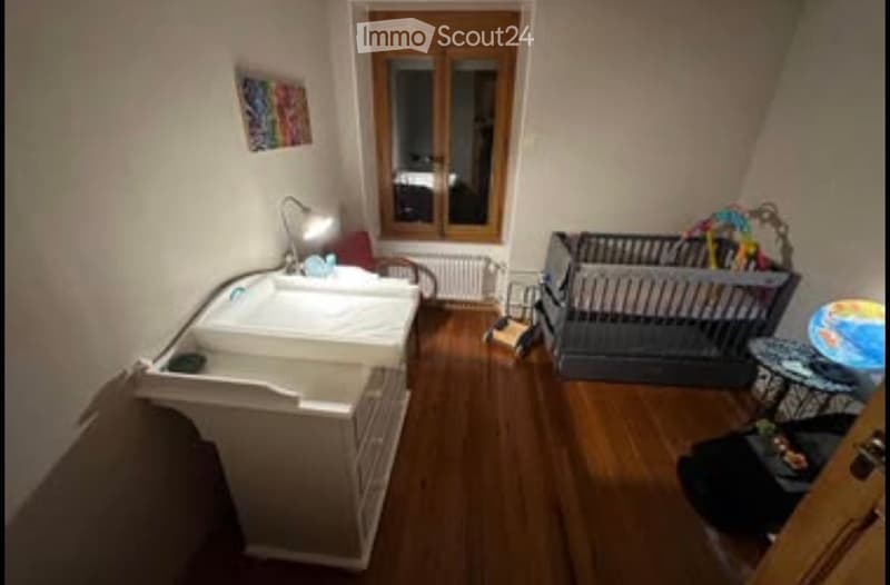 A small, cozy bedroom with a single bed, a small table with a lamp, a chair, a crib, and various toys scattered around. The room has wooden flooring and a window with wooden frames.