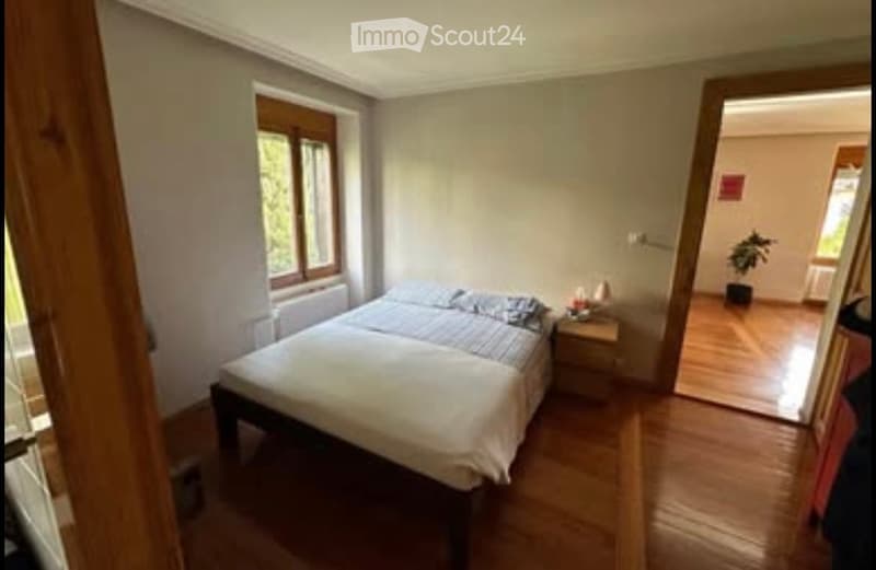 Cozy bedroom with wooden flooring, a double bed with pillows and blankets, a nightstand with a lamp, and a window with wooden frames. A small plant is visible in the adjacent room.