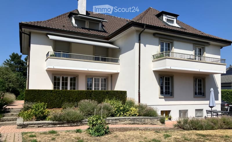 Two-story house with white exterior, multiple windows, balconies, and a well-maintained garden with shrubs and a patio area.