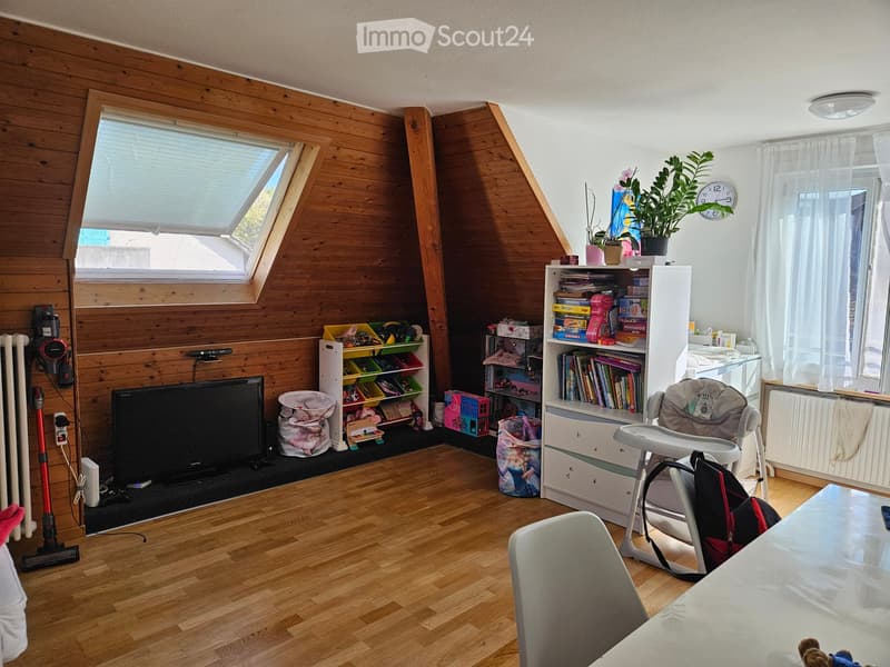 A well-lit attic room with wooden walls and flooring, featuring a large window, a television, a bookshelf, a baby chair, and a table with a chair.