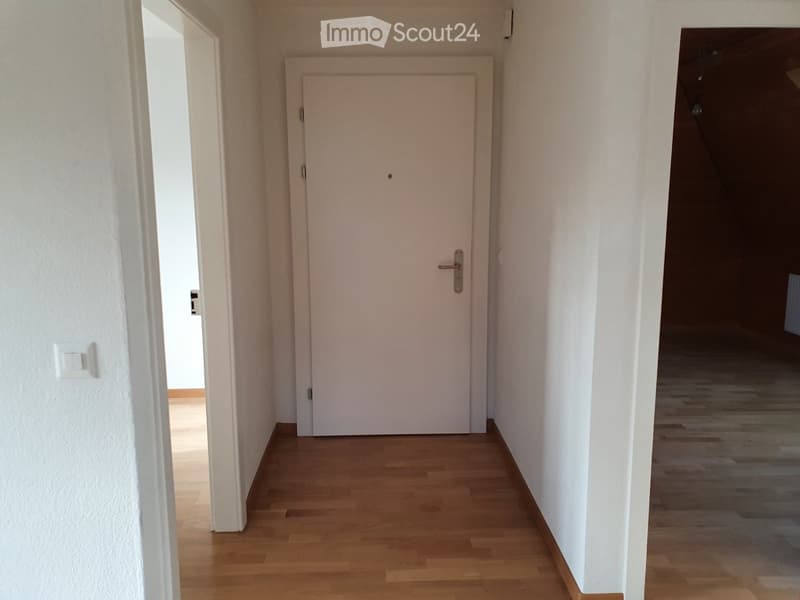A narrow hallway with light wooden flooring and white walls, featuring a closed white door and an open doorway leading to another room.