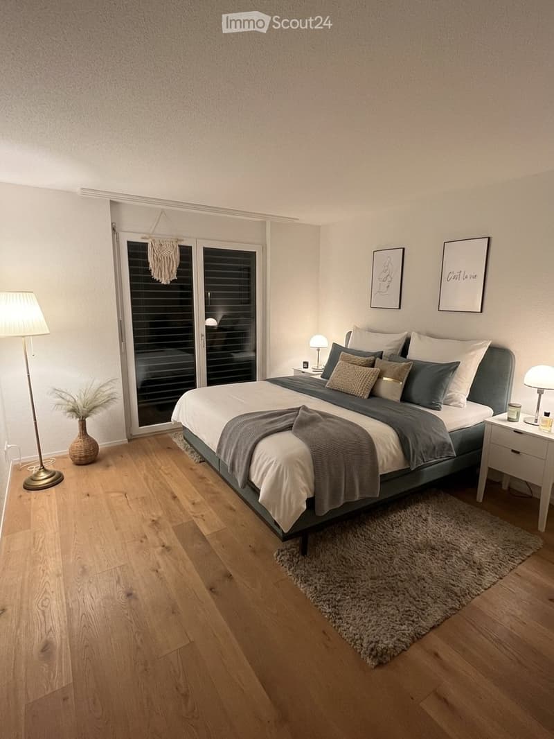 Modern bedroom with wooden flooring, a large bed with pillows and blankets, nightstands with lamps, a rug, a decorative vase, and picture frames on the wall.