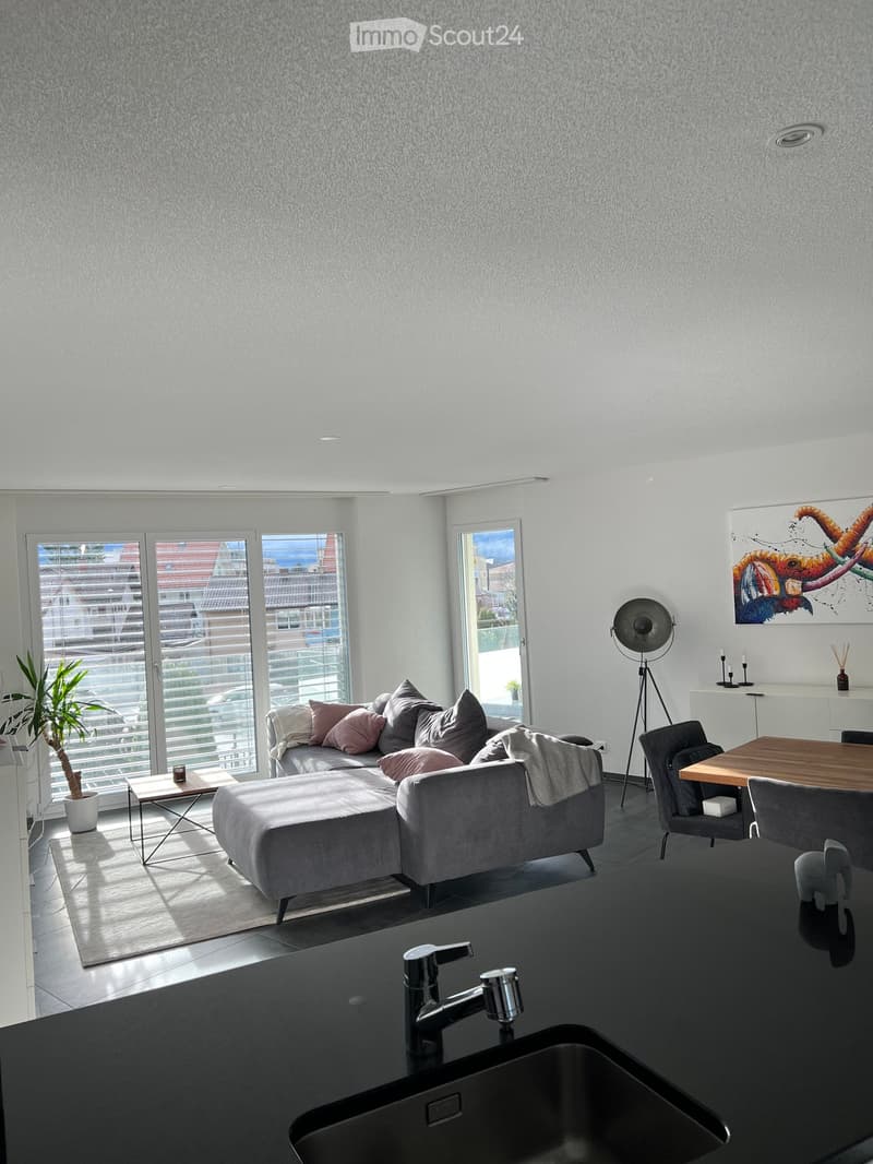 Modern living room with grey sofa, ottoman, coffee table, dining area, and large windows offering a view of the city.