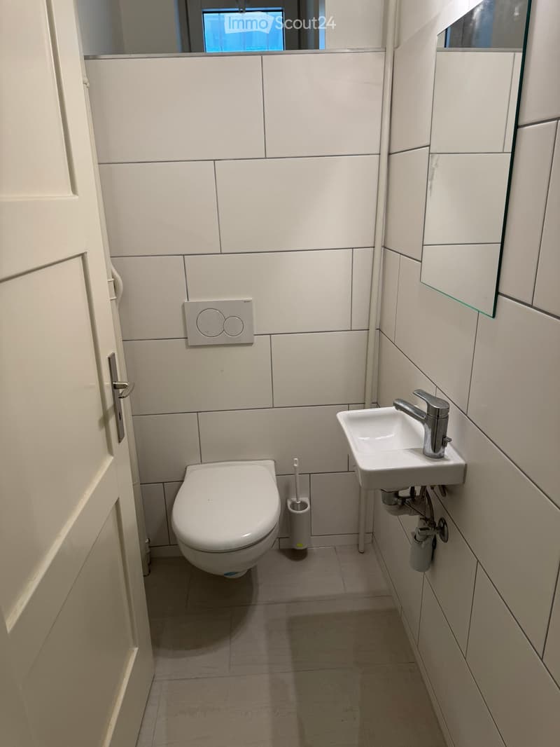 Modern bathroom with white tiles, a toilet, a sink with a chrome faucet, and a toothbrush holder. The space is compact with a large mirror above the sink.