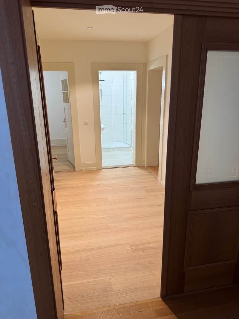 Empty hallway with wooden flooring, light-colored walls, and open doors leading to a bathroom and another room.