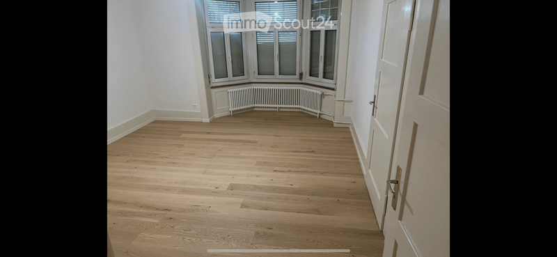 Empty room with light wooden flooring, white walls, large windows with blinds, and a radiator below the windows.