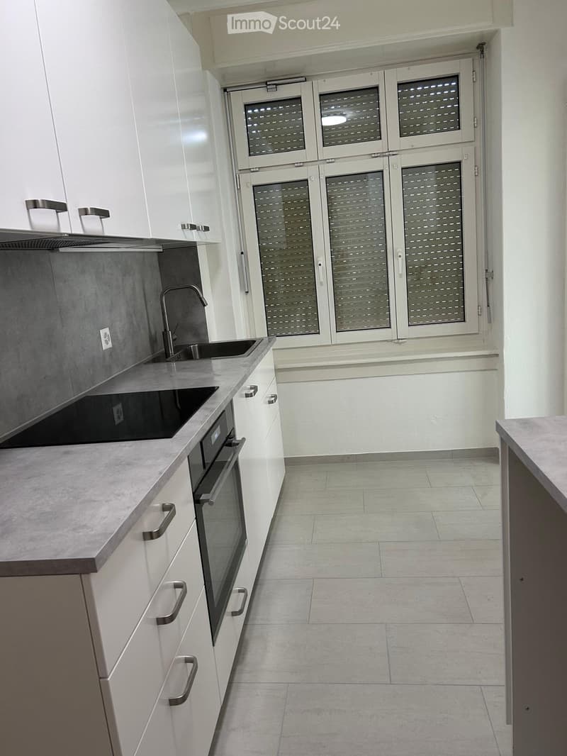 Modern kitchen with white cabinets, stainless steel sink, and built-in oven. Grey countertops and tiled floor.