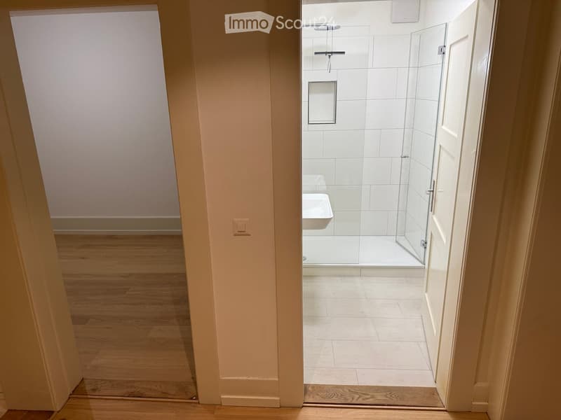 Modern bathroom with white tiles, glass shower enclosure, and a sink visible through an open doorway.