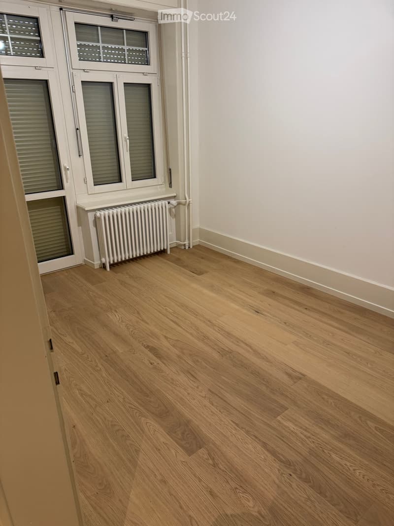 Empty room with light wooden flooring, white walls, and a radiator near a window with blinds.