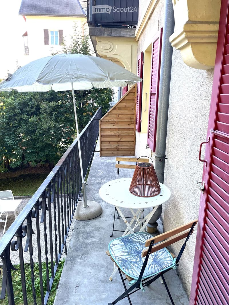 Balcony with a small round table, a wicker lamp, a folding chair with a blue patterned cushion, and an umbrella.
