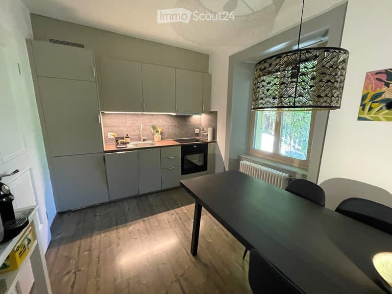 Modern kitchen with grey cabinets, a sink, oven, and a dining area with a table and chairs. A large window provides natural light, and a decorative lamp hangs from the ceiling.