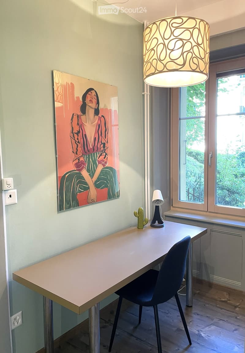 A modern, well-lit room with a large wooden table, a black chair, a hanging lamp, a small cactus, and a colorful painting on the wall. The room has large windows with a view of greenery outside.