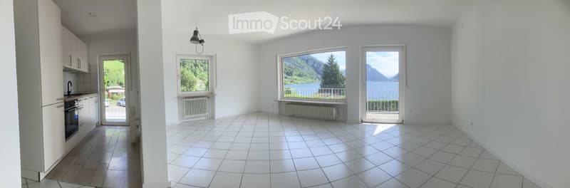 Bright, unfurnished living space with large windows offering scenic views, tiled floor, and a small kitchen area with cabinets and appliances.