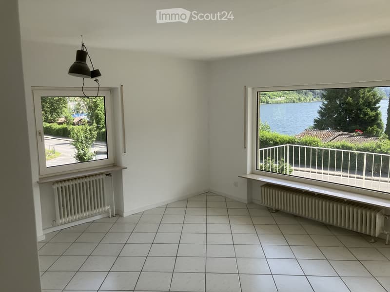 Empty room with white tiled flooring, white walls, and large windows offering scenic views of a lake and surrounding greenery.
