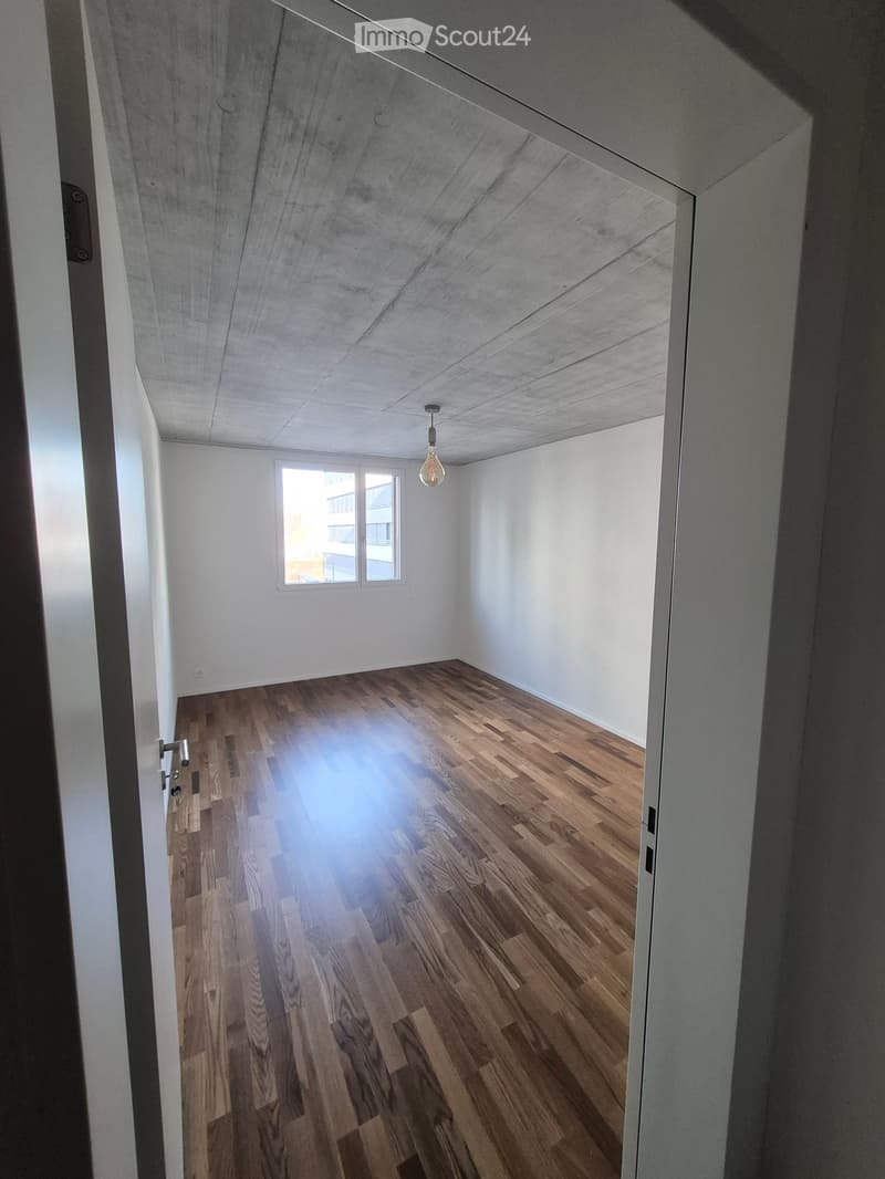 Empty room with wooden flooring, white walls, and a single window letting in natural light. The ceiling has a modern, textured finish with a single hanging light fixture.