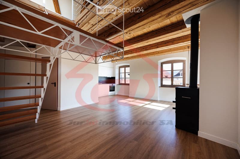 Bright, open-plan space with wooden flooring, exposed wooden beams, and large windows. Features a staircase leading to an upper level, a modern kitchen area, and a wood-burning stove.