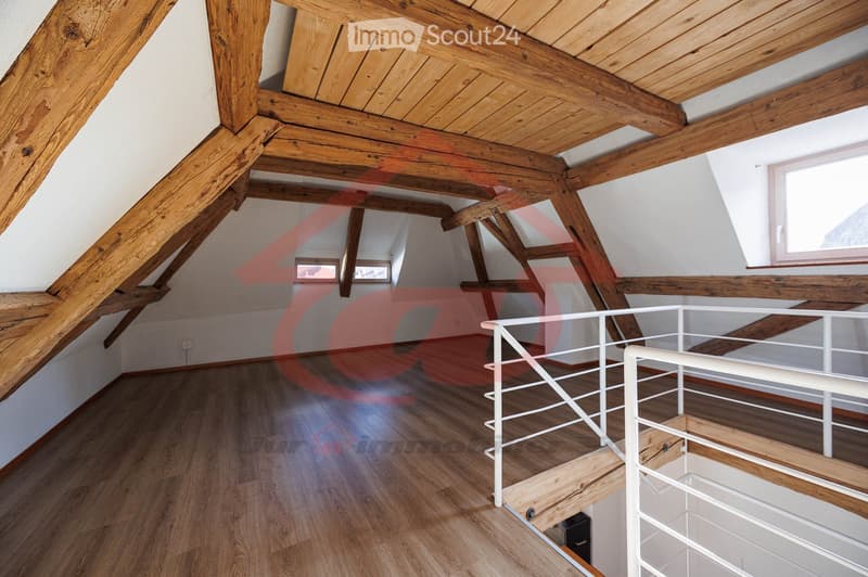 An attic space with exposed wooden beams and a wooden floor, featuring a white metal railing staircase leading to a lower level. The room has white walls and several windows allowing natural light to enter.