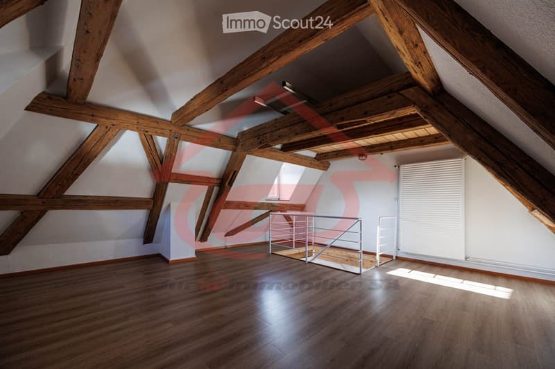An empty attic space with exposed wooden beams, wooden flooring, and a small metal staircase leading to a loft area. The room has white walls and a skylight window allowing natural light to enter.