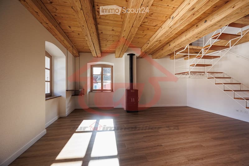 Empty room with wooden floor, exposed wooden ceiling beams, white walls, and a staircase leading upwards. Features include a window with a wooden frame, a chimney, and a small built-in shelf on the left side.
