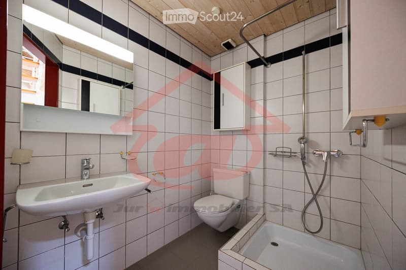 Bathroom with white tiles, a sink, toilet, bathtub, mirror, and a small cabinet. The room features a wooden ceiling and a window providing natural light.
