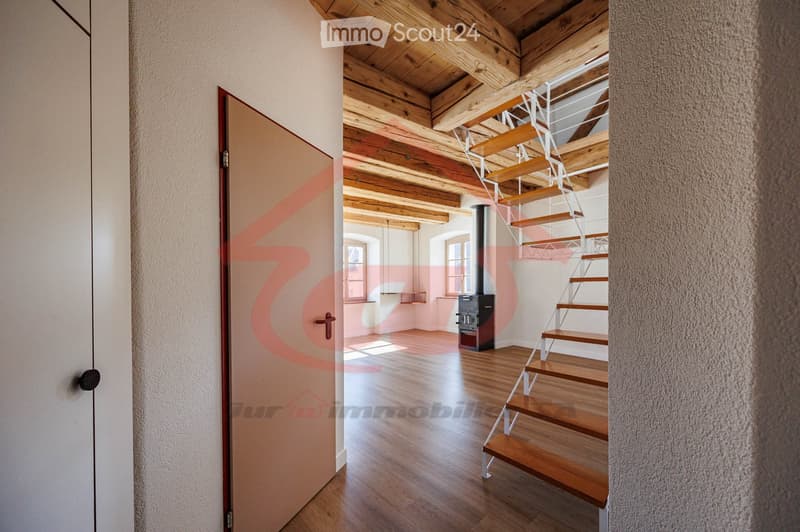A modern hallway with wooden flooring, exposed wooden beams on the ceiling, a staircase leading upwards, and a visible fireplace in the background.
