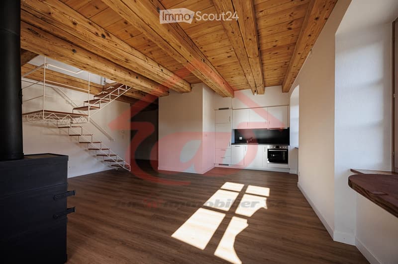 Open-plan space with wooden floor and ceiling, staircase leading to an upper level, modern kitchen area with cabinets and oven, and a large black wood-burning stove.