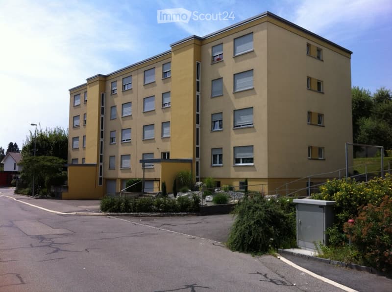 Four-story yellow apartment building with multiple windows and a small landscaped area in front.
