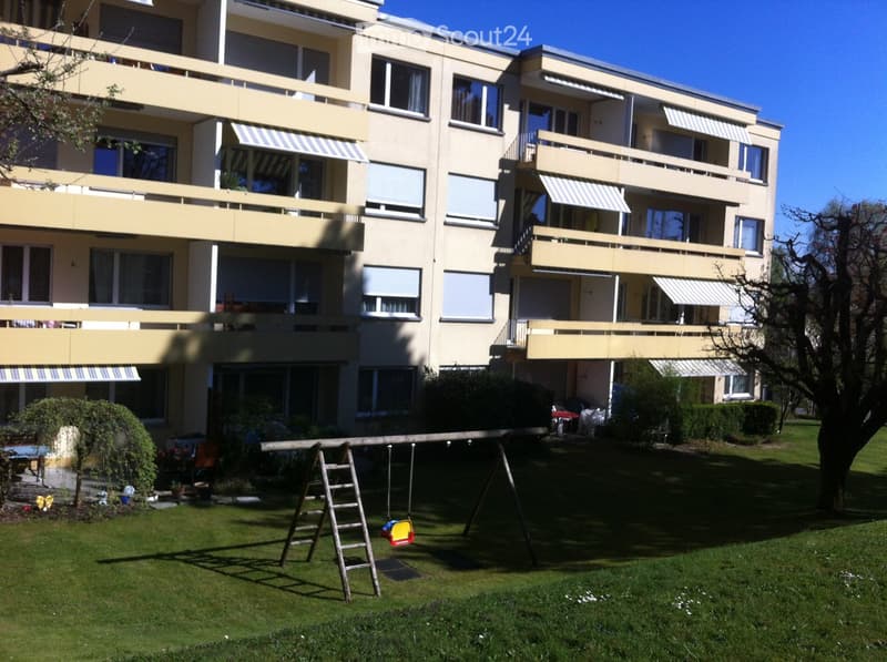 Multi-story residential building with balconies, large windows, and striped awnings. The surrounding area includes a grassy lawn, a swing set, and some trees.